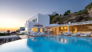 Un espace moderne aménagé au bord de la piscine, avec des chaises longues, un bar et une vue sur la mer au coucher du soleil.