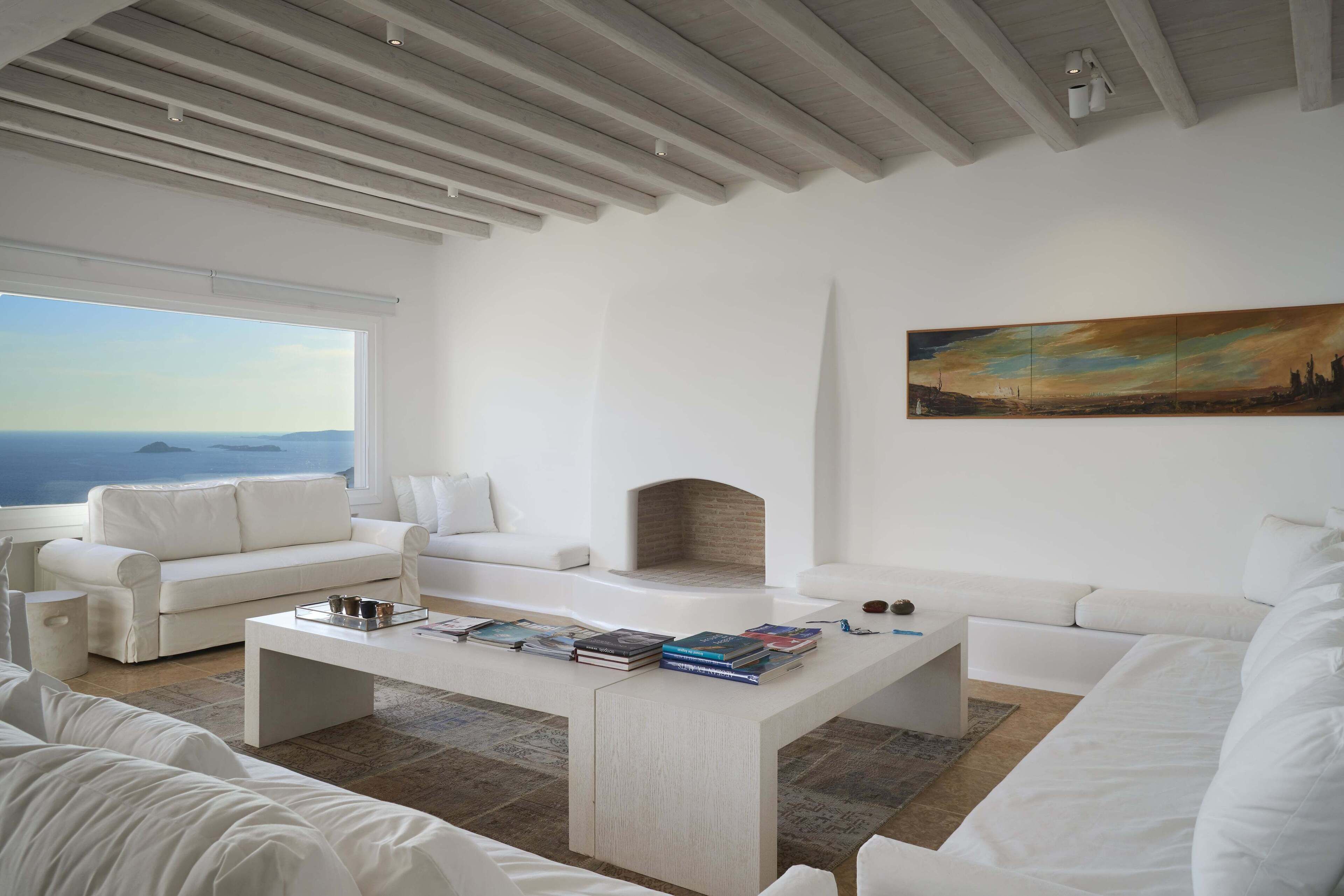 A modern living room with white furniture, a fireplace, and a large window with a view of the sea.