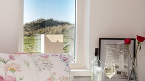 Un dormitorio con cama, ventana con vista a una colina, mesita de noche con una botella, un vaso y un florero con una flor.