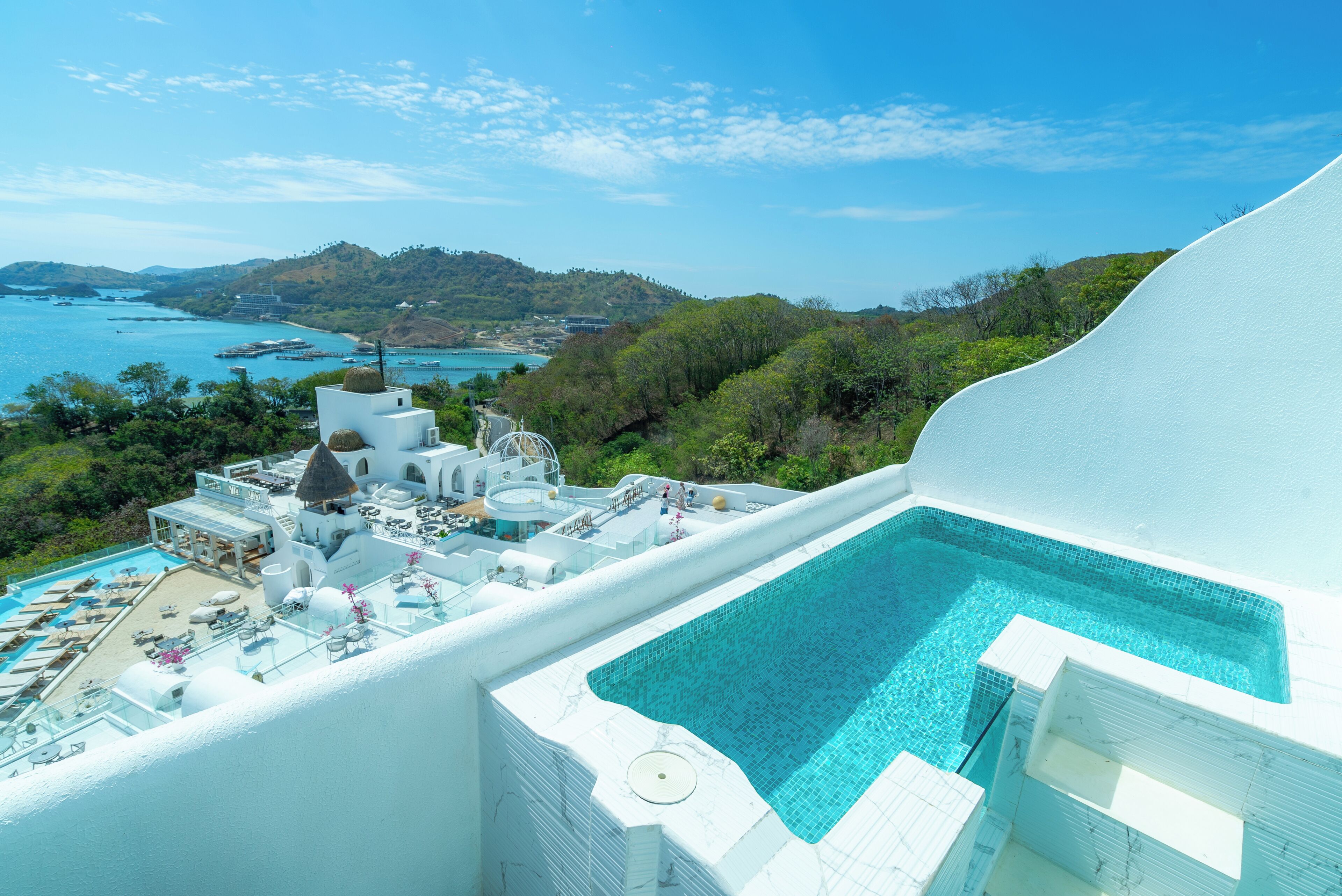 A resort with a large pool, a beach area, and a view of the sea and hills.