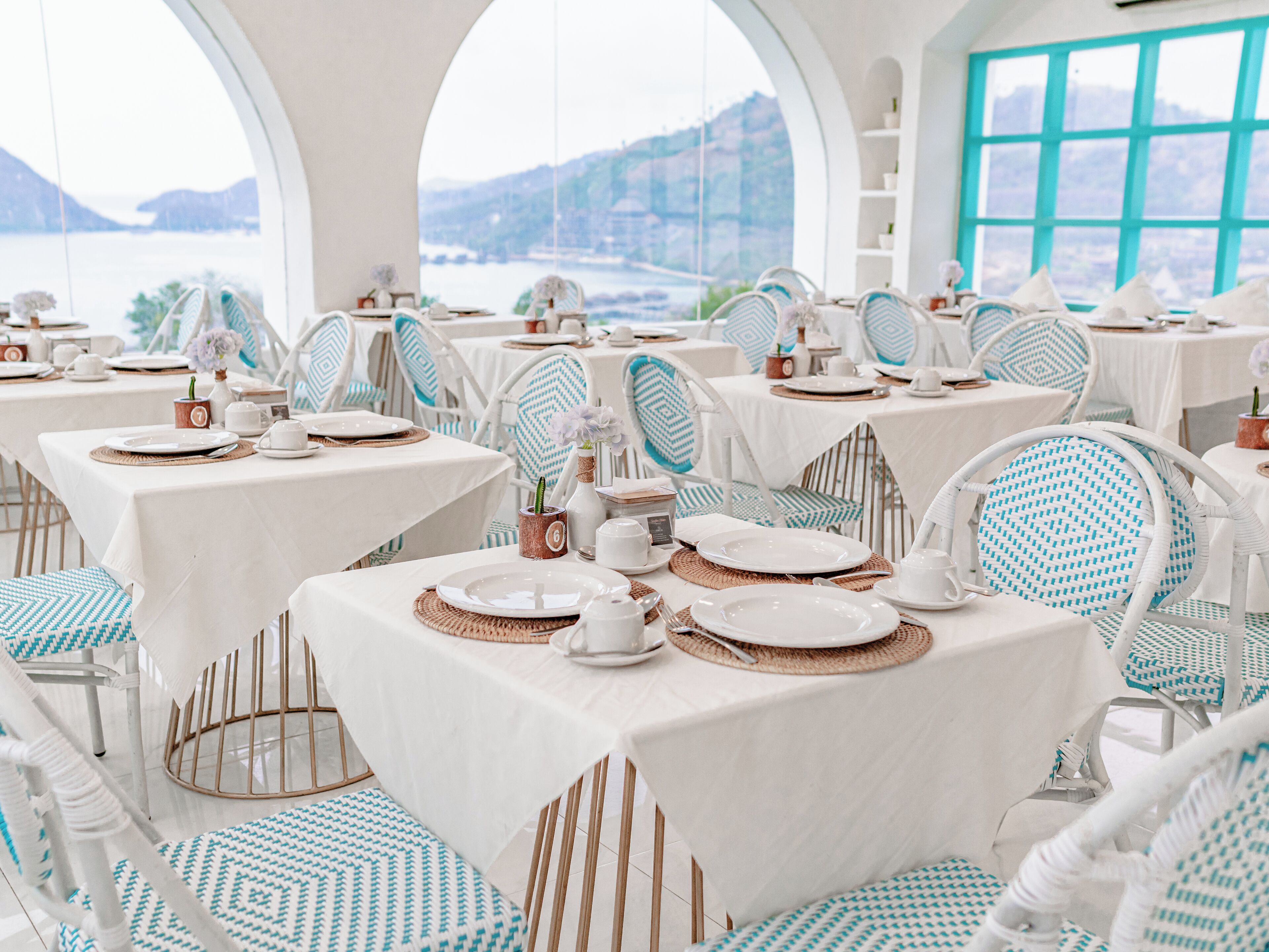 Un comedor con mesas dispuestas para una comida, sillas con cojines estampados y vistas al mar a través de amplios ventanales.