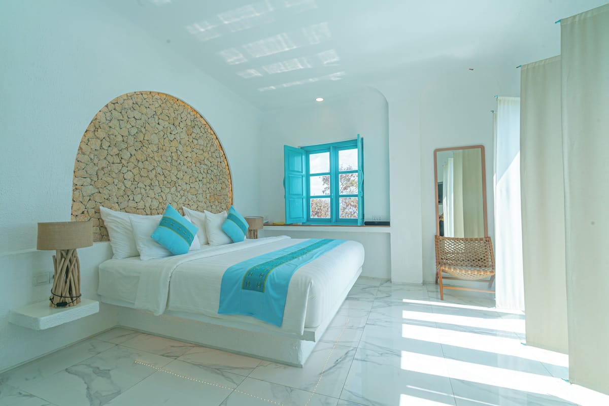 A bedroom with a large bed, a window with blue shutters, a mirror, and a wicker chair.