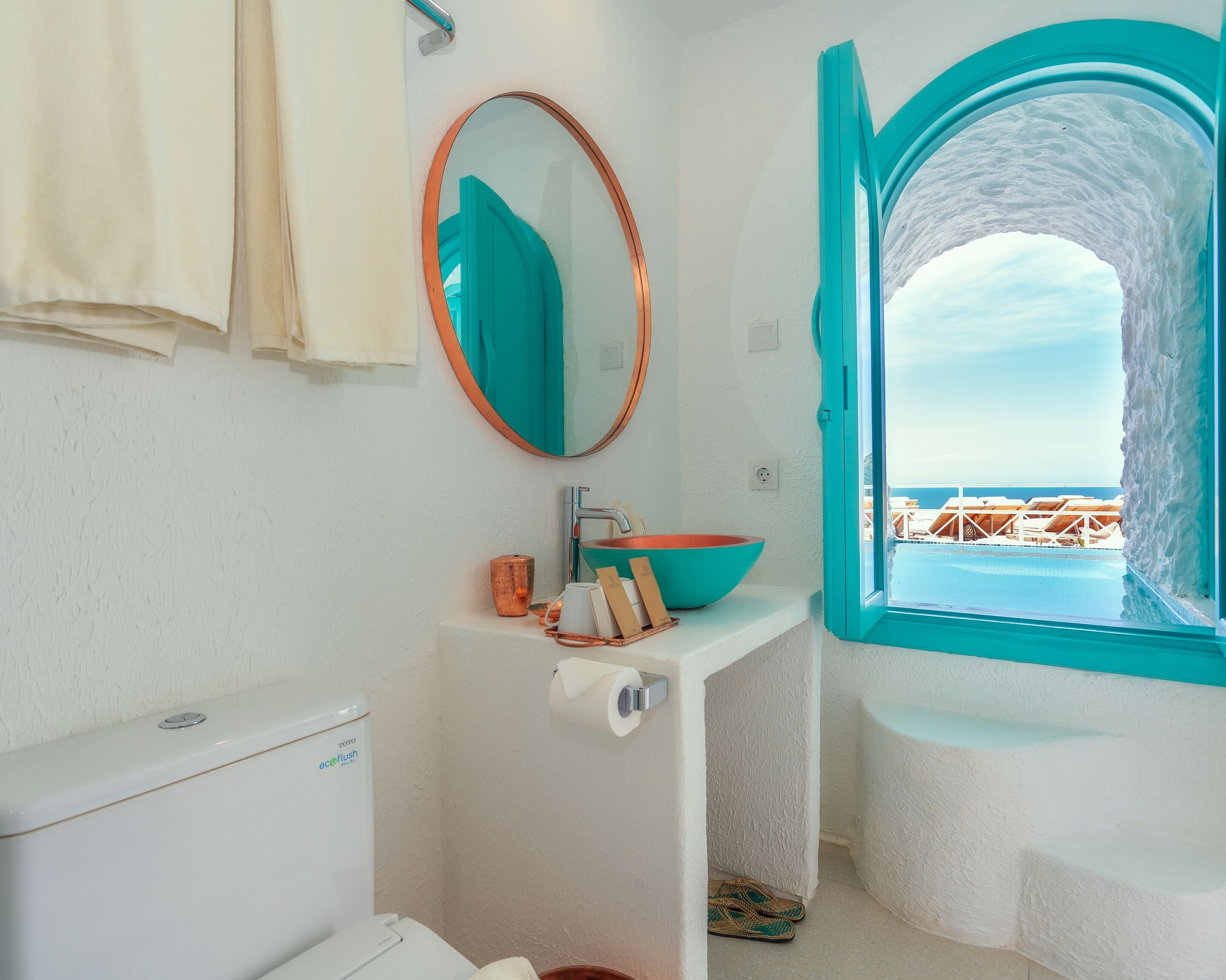 A bathroom with a white sink, a round mirror, and a view of the sea through a turquoise arch window.