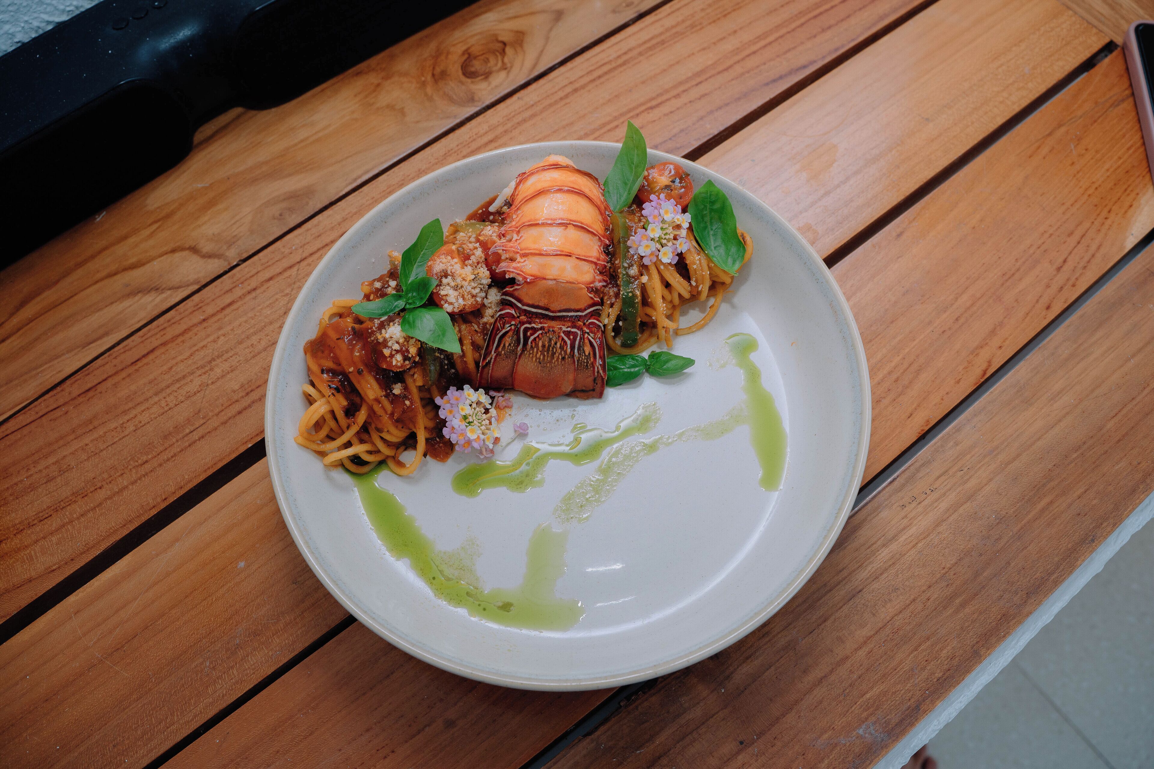 A plate of pasta with a lobster tail, garnished with basil and flowers, on a wooden table.