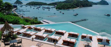Un espace aménagé au bord de la piscine, avec des chaises longues et des tables, une vue dégagée sur la mer et les bateaux, et une île boisée en arrière-plan.