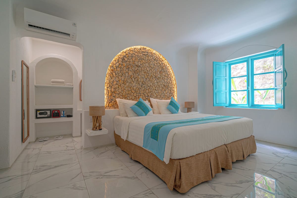A bedroom with a large bed, a stone wall, a window with blue shutters, and a small shelf with a radio and books.