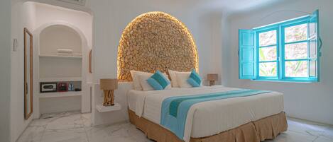 A bedroom with a large bed, a stone wall, a window with blue shutters, and a small shelf with a radio and books.