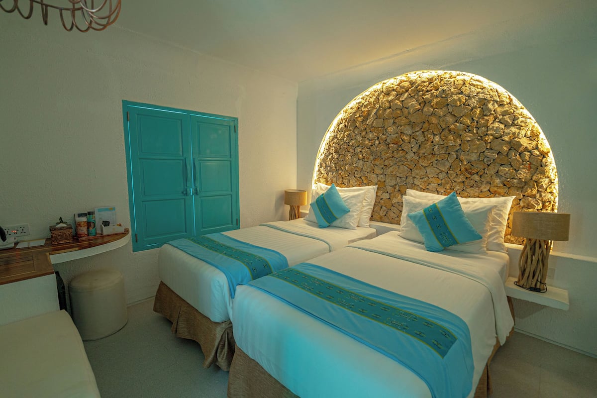 A hotel room with two beds, a blue wardrobe, a stone wall, and a small desk with a lamp.