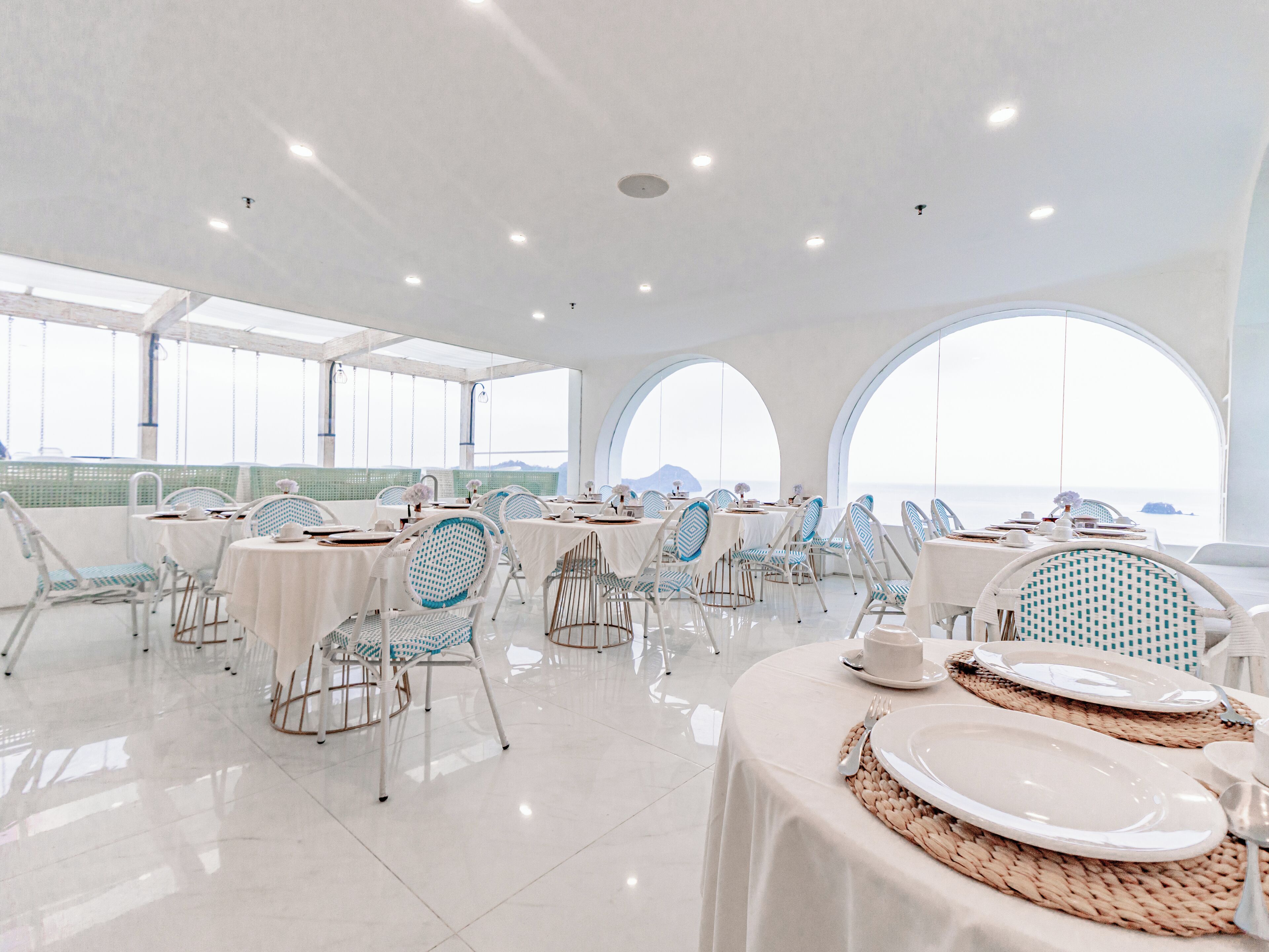 A spacious dining area with white tables and chairs, large arched windows, and a view of the sea.
