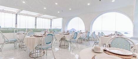 A spacious dining area with white tables and chairs, large arched windows, and a view of the sea.