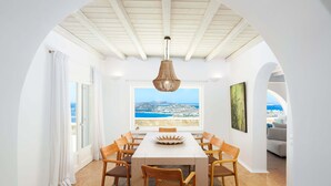 Un comedor con mesa y sillas, vista al mar a través de una ventana y un techo con vigas de madera.