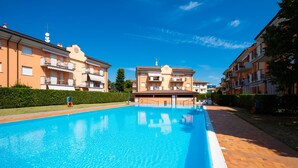 A swimming pool with a clear blue water surface, surrounded by apartment buildings with balconies and a hedge in the foreground. - Appartamento Francesca (Sirmione)