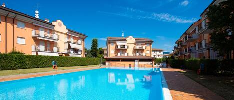 A swimming pool with a clear blue water surface, surrounded by apartment buildings with balconies and a hedge in the foreground.