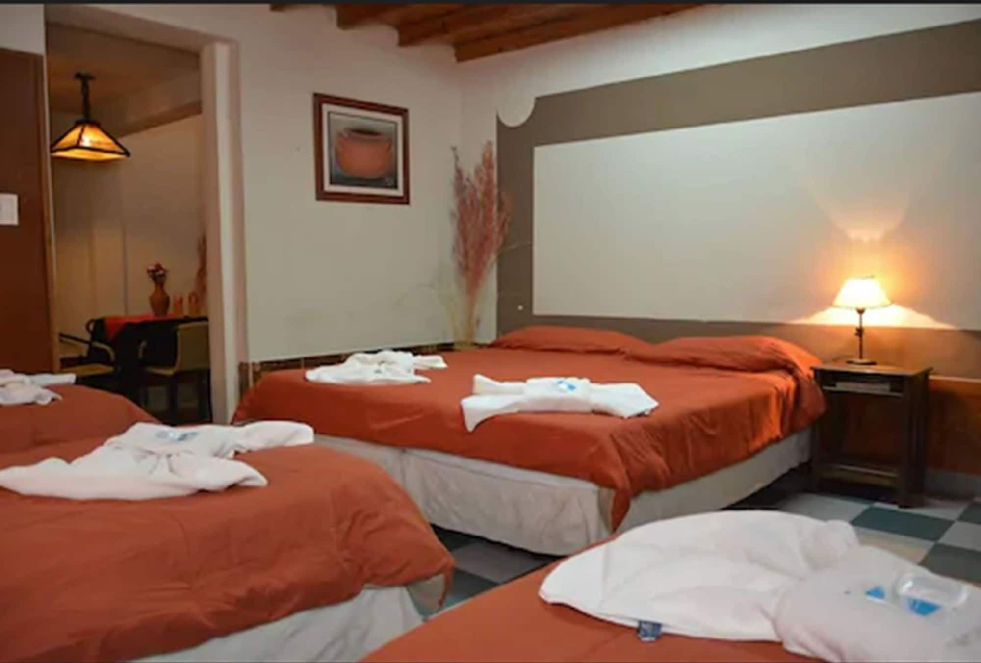 Une chambre d’hôtel avec trois lits, chacun recouvert d’une couverture orange et agrémenté de serviettes blanches. On y trouve une petite table avec une lampe, un tableau encadré au mur et un vase contenant des plantes séchées.
