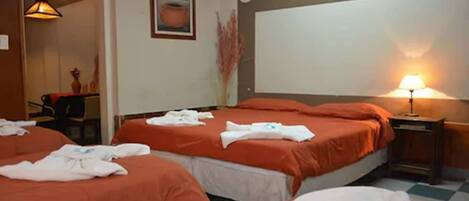 Une chambre d’hôtel avec trois lits, chacun recouvert d’une couverture orange et agrémenté de serviettes blanches. On y trouve une petite table avec une lampe, un tableau encadré au mur et un vase contenant des plantes séchées.