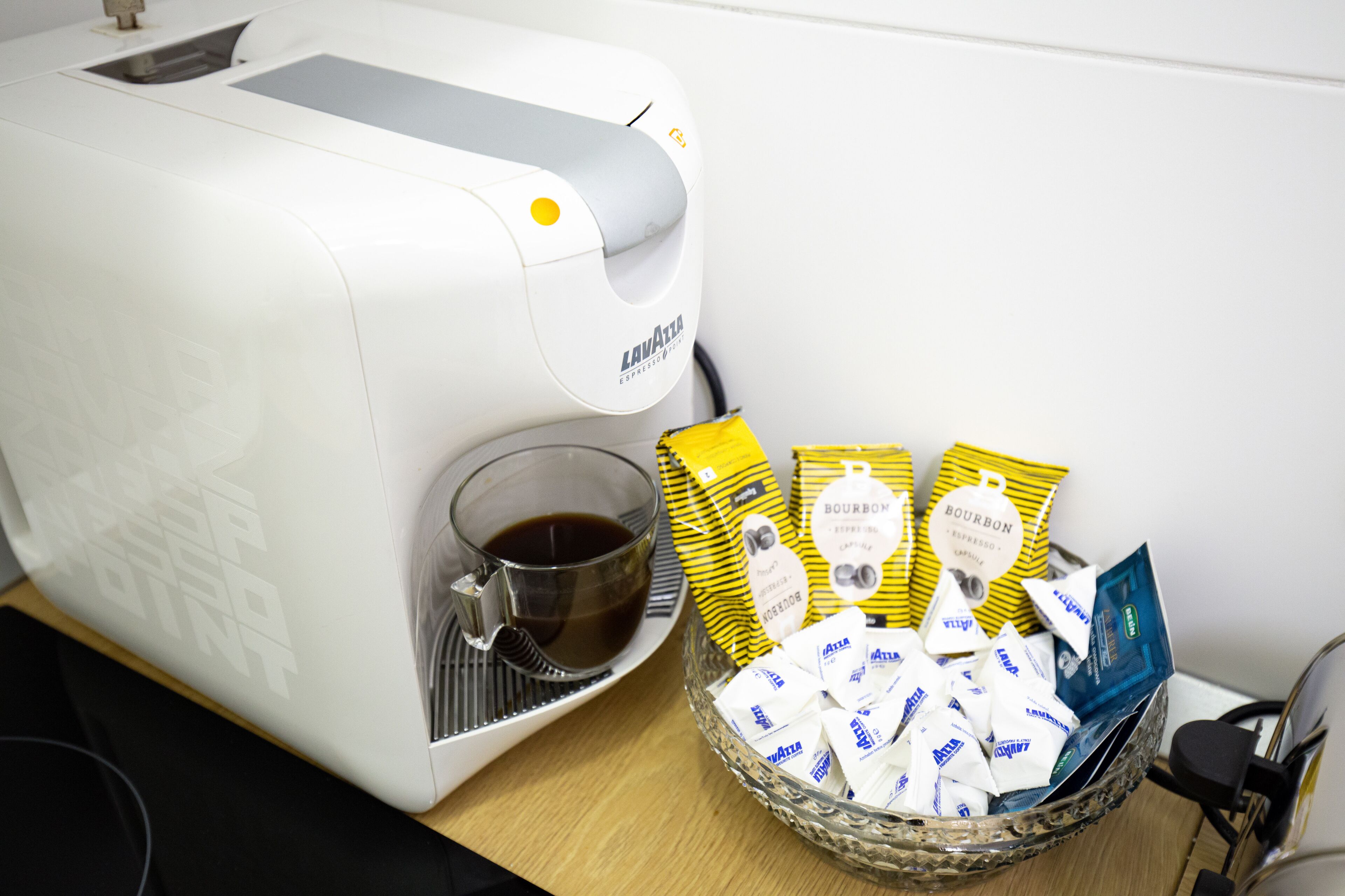Een koffieapparaat schenkt koffie in een kopje, een schaal met koffiepads en een glas koffie op een tafel.
