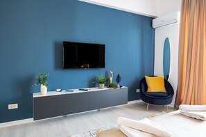 Une chambre moderne avec un mur bleu foncé, un téléviseur fixé au mur, une console grise, une chaise jaune et un lit avec du linge de lit blanc.