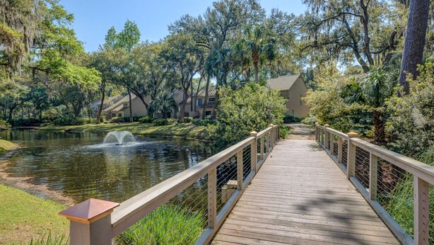 A wooden boardwalk leading to a body of water with a fountain, surrounded by lush greenery and trees.