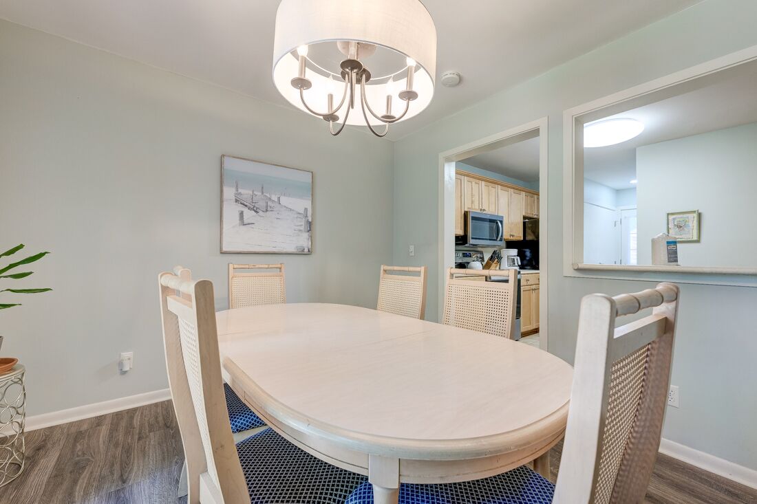 Ein Essbereich mit einem runden Tisch, vier Stühlen, einer Chandelierleuchtre, einem gerahmten Bild an der Wand und einer durch einen offenen Durchgang einsehbarer Küche.