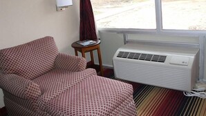 A room with a patterned sofa, a small table with a book, a white air conditioner, and a window with a view of the outside. - Ramada Perry near Fairgrounds (Perry)