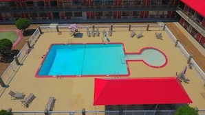 A swimming pool with two sections, surrounded by lounge chairs and umbrellas, with a red-roofed building in the background. - Ramada Perry near Fairgrounds (Perry)
