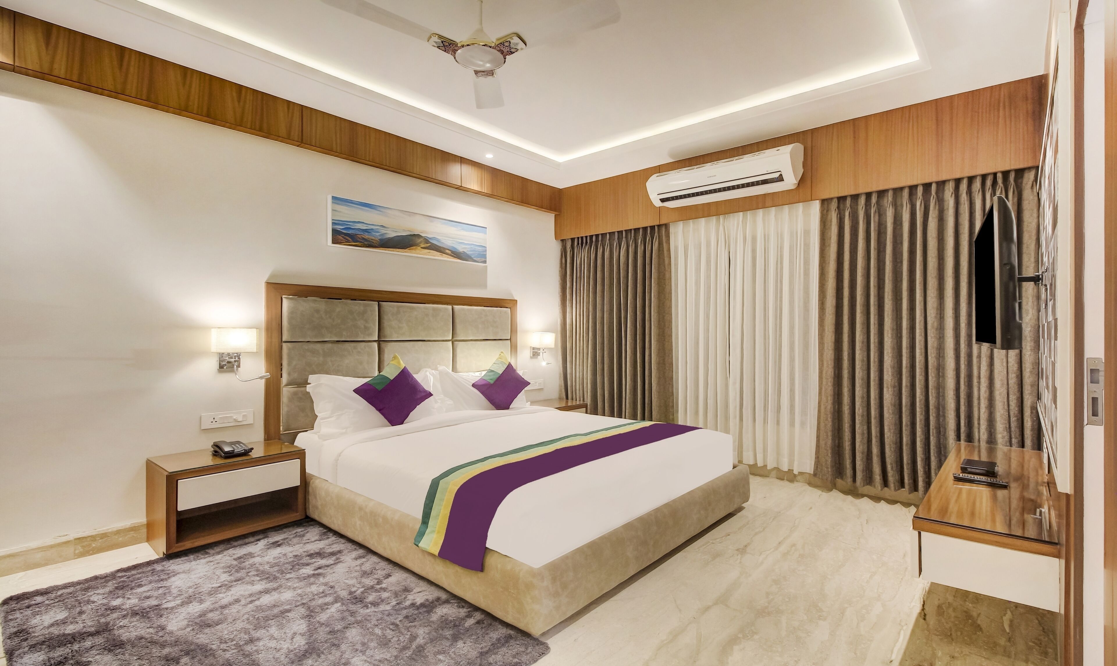 Una camera d'hotel moderna con un letto grande, pannelli in legno, un condizionatore d'aria, una ventola a soffitto e un quadro appeso al muro.