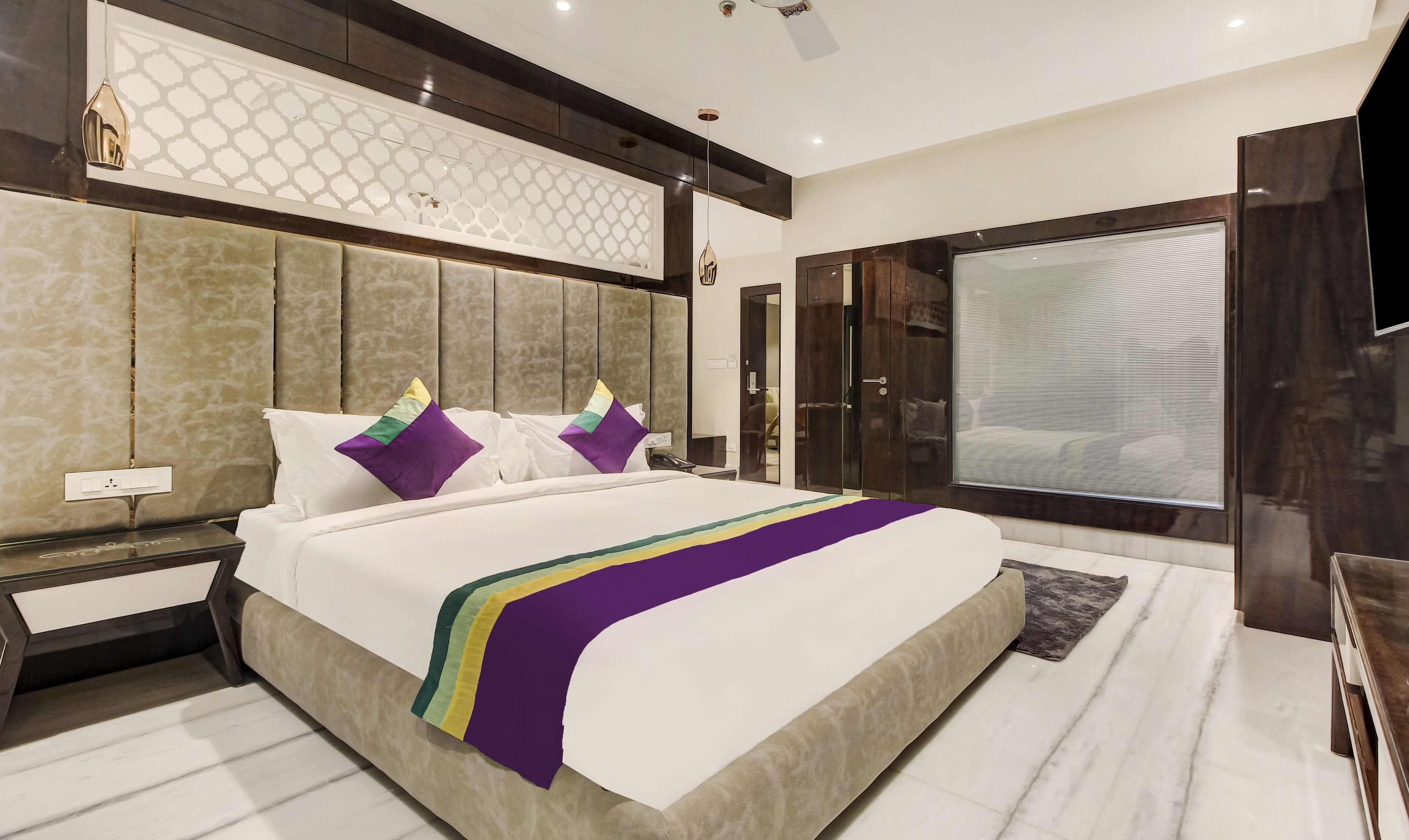 A modern hotel room with a large bed, a nightstand, a desk, and a large window with blinds.