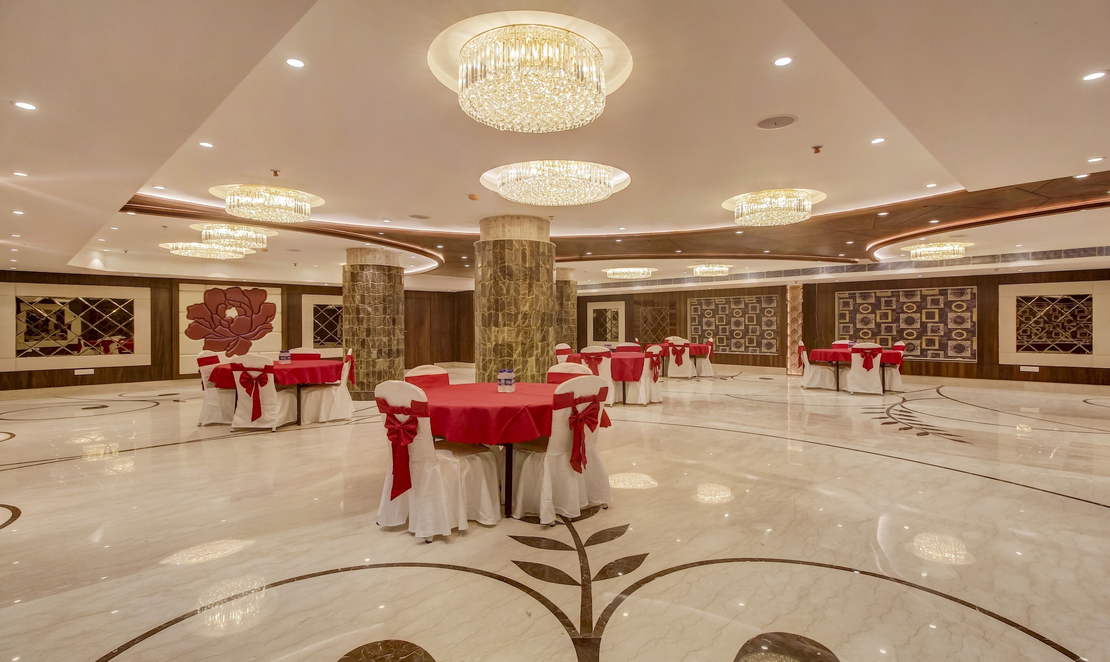 Una amplia sala de banquetes con múltiples mesas redondas, cada una cubierta con manteles rojos y sillas blancas decoradas con lazadas rojas. La sala cuenta con suelos de mármol con motivos florales, grandes columnas decorativas y lujosos candelabros.