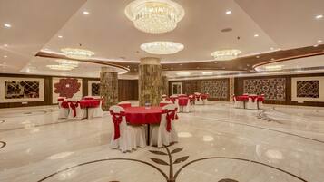 Una amplia sala de banquetes con múltiples mesas redondas, cada una cubierta con manteles rojos y sillas blancas decoradas con lazadas rojas. La sala cuenta con suelos de mármol con motivos florales, grandes columnas decorativas y lujosos candelabros.