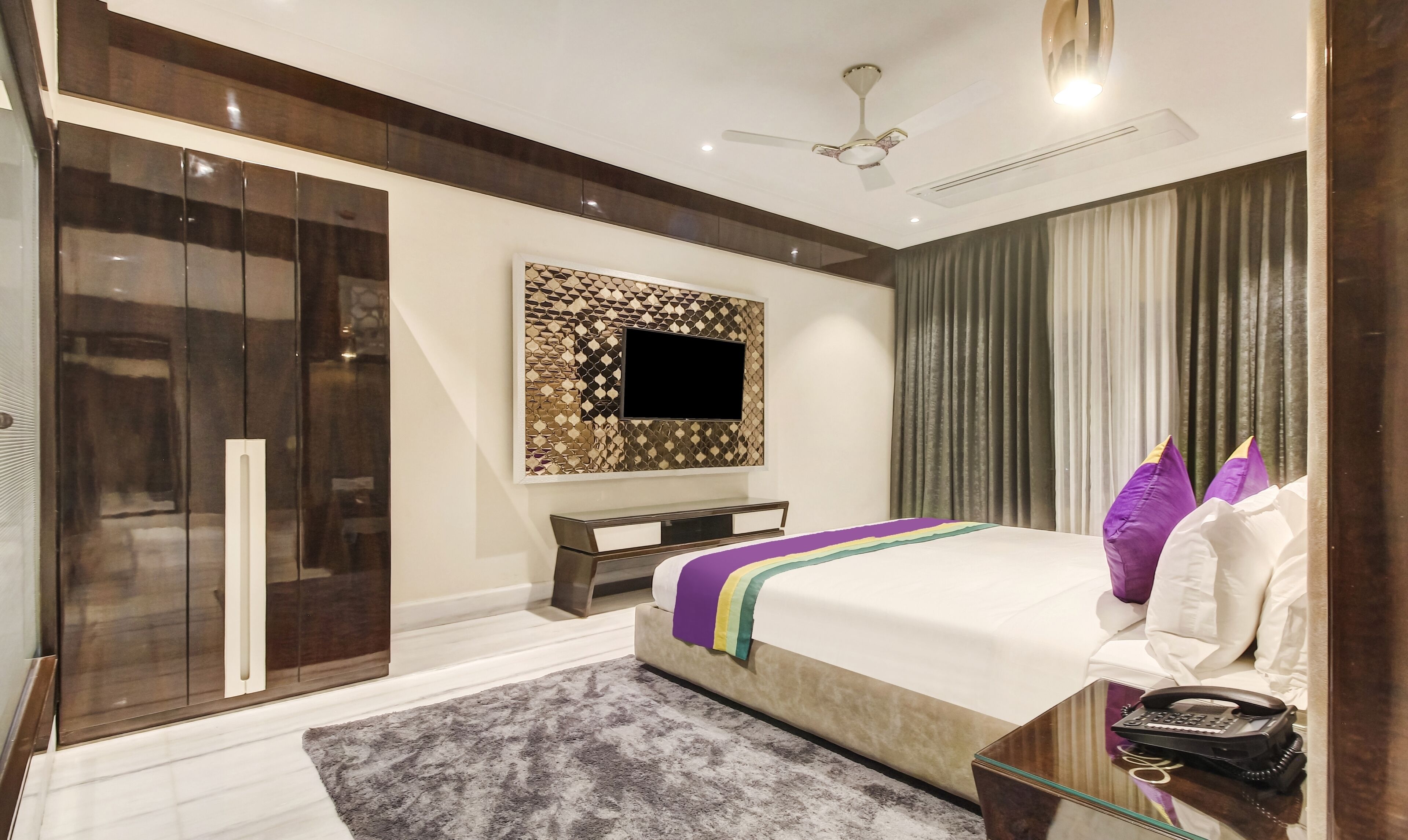 A modern bedroom with a large bed, a TV mounted on a decorative wall, a wardrobe, and a bedside table with a phone.