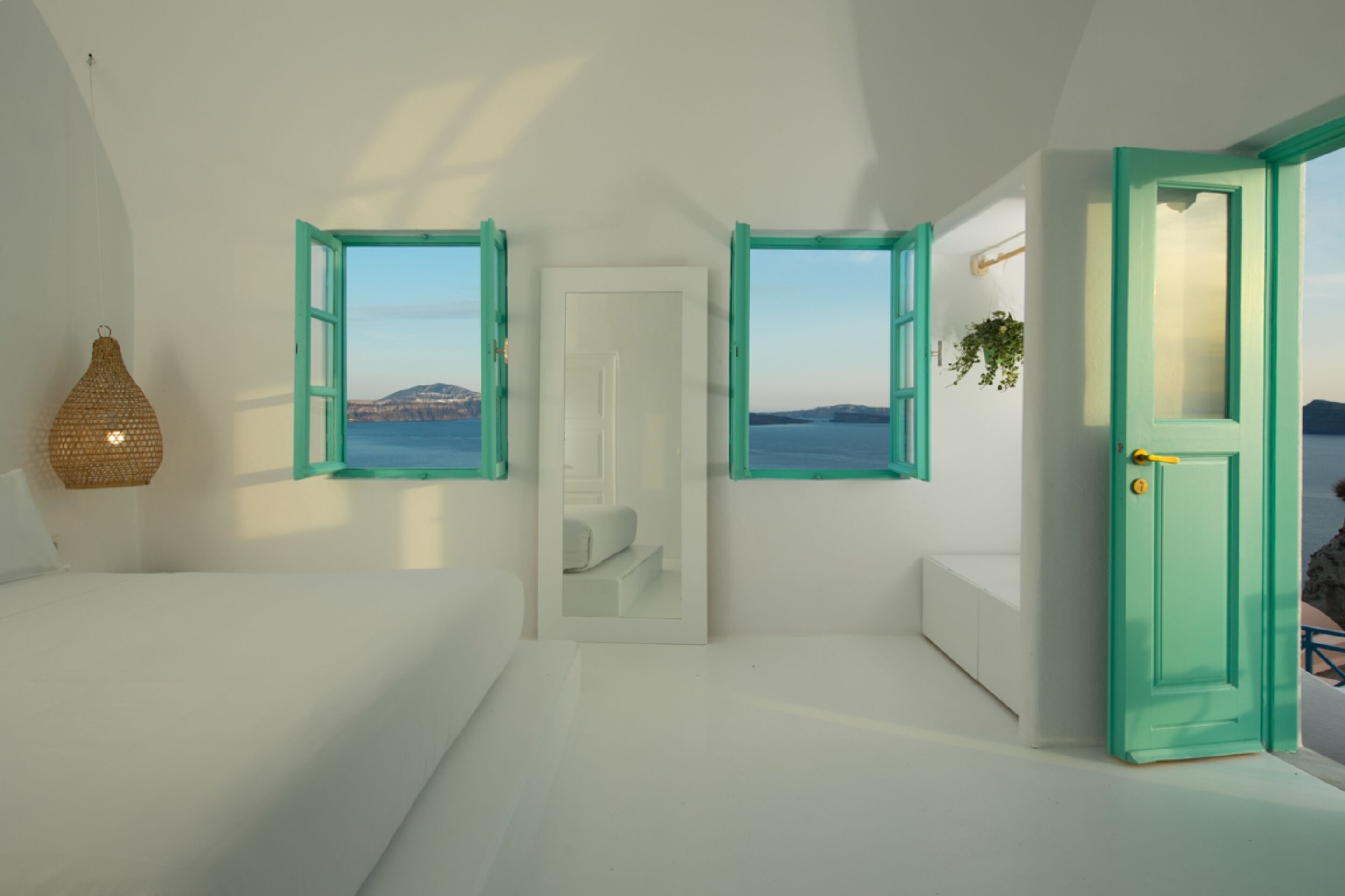 Une chambre avec un lit, un luminaire suspendu et deux fenêtres aux cadres turquoise, offrant une vue sur la mer.