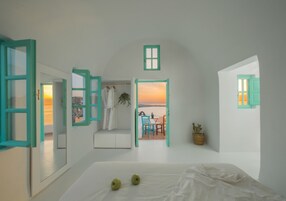 Una habitación con una cama, dos ventanas con marco verde, vistas al atardecer y una mesa con sillas afuera.