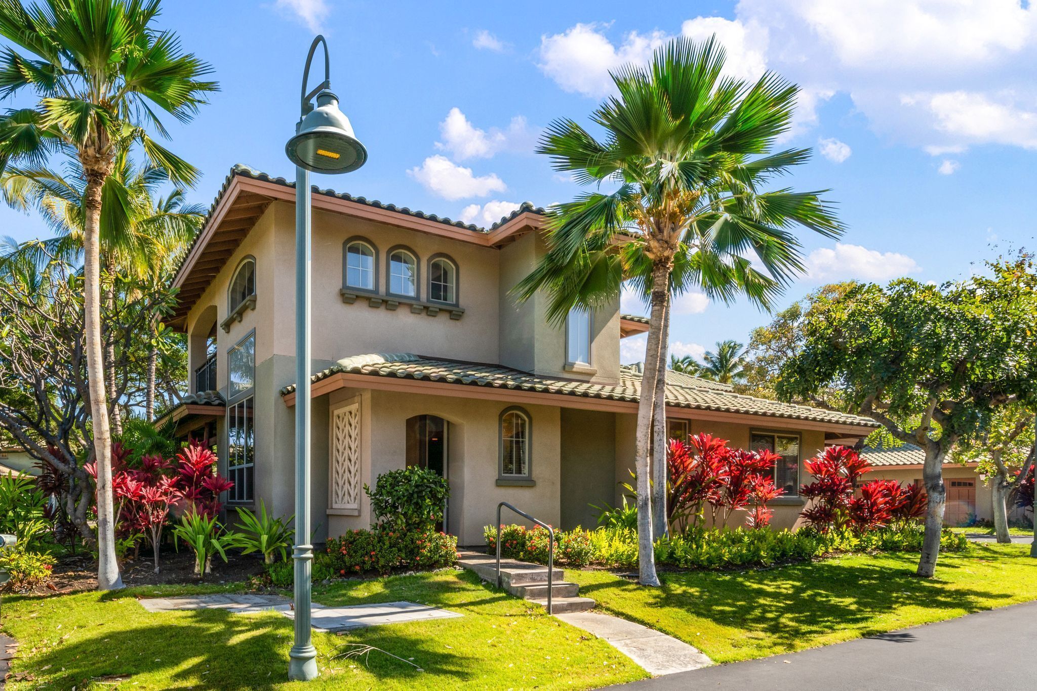 A two-story house with a tiled roof, a front porch, and a well-maintained garden with palm trees and red flowering plants.