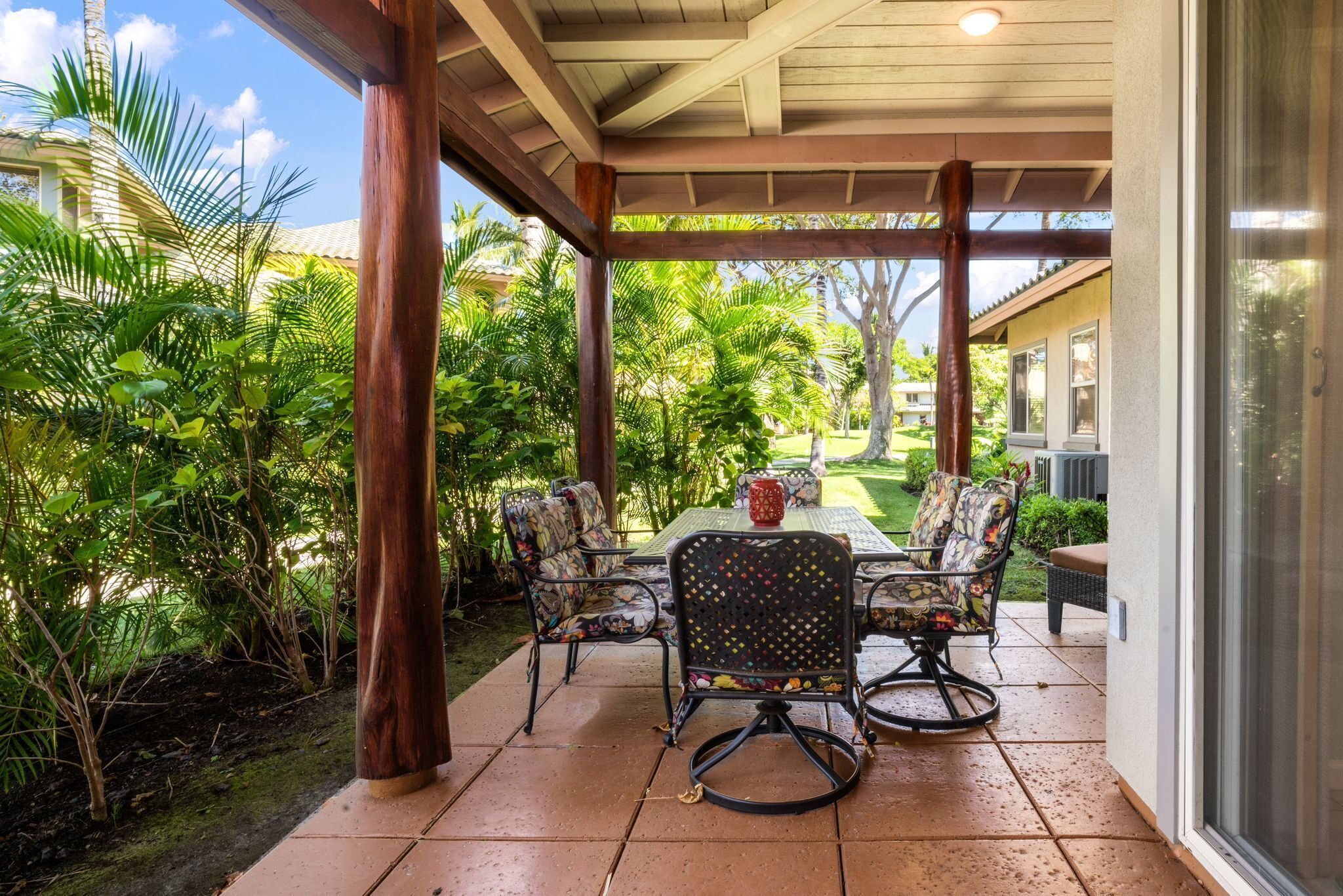 A covered patio with a table and chairs, surrounded by lush greenery and a view of a residential area.