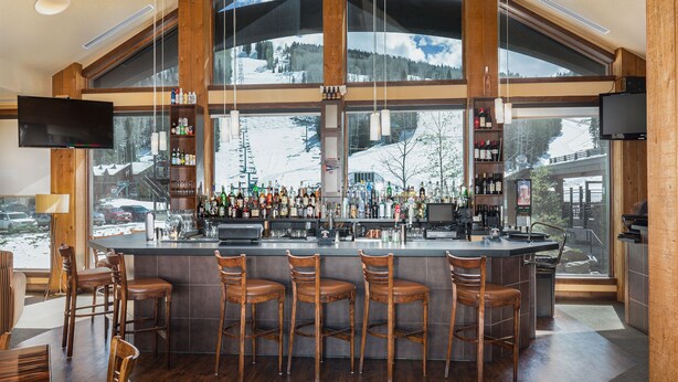Un espace bar avec une grande fenêtre donnant sur une montagne enneigée, des tabourets de bar en bois et une variété de bouteilles et de verres exposés sur des étagères.
