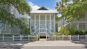 Une maison à deux étages, dotée d’un balcon et d’une véranda, entourée d’une clôture en bois peinte en blanc.