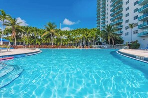 A large, clear swimming pool surrounded by palm trees and a tall building with balconies.