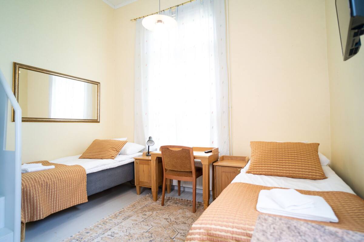 Une chambre d’hôtel avec deux lits simples, un bureau, une chaise, un miroir et une fenêtre avec des rideaux.