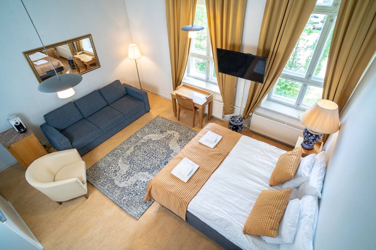 Une chambre d’hôtel avec un lit, un canapé, un bureau, une chaise, une télévision et une fenêtre avec des rideaux.