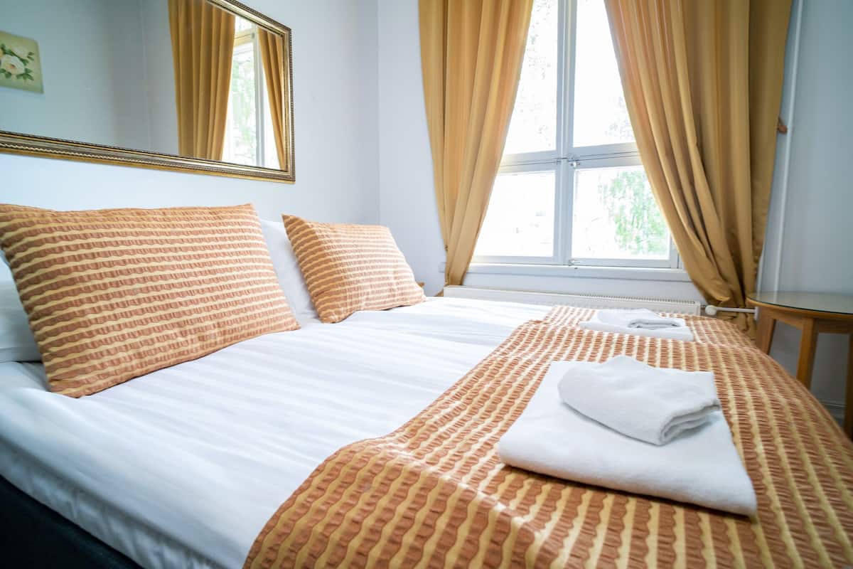Un lit bien fait, avec une couvre-lit à rayures et des coussins assortis, une fenêtre avec des rideaux et une petite table accompagnée d’une chaise.