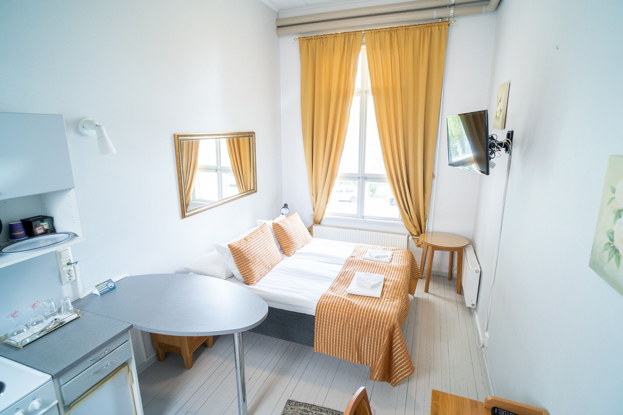 A compact hotel room with a bed, a small table, a chair, a desk, and a window with curtains.