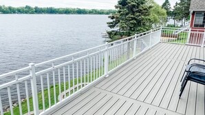 A wooden deck with a white railing overlooking a lake, a tree, and a building with a red roof.