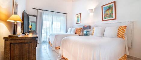 Habitación de hotel con dos camas, un aparador de madera, un escritorio con lámpara y un ventanal con cortinas translúcidas.