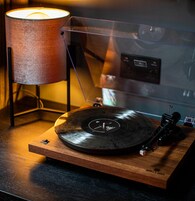 Tocadiscos sobre un soporte de madera con una cubierta protectora transparente, una lámpara y un reproductor de discos en el fondo.
