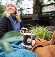 Una persona sosteniendo una taza con un diseño de calavera, otra persona al fondo sosteniendo una taza, y una planta en primer plano.