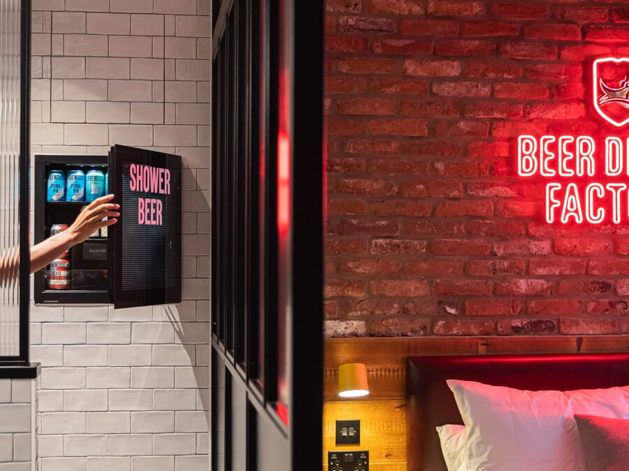A hotel room with a brick wall, a neon sign reading "BEER FACT", and a minibar with a "SHOWER BEER" sign.