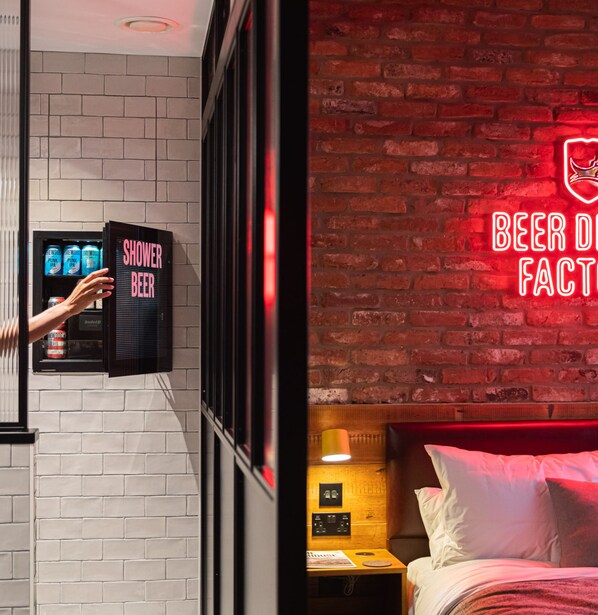 A hotel room with a brick wall, a neon sign reading "BEER FACT", and a minibar with a "SHOWER BEER" sign.