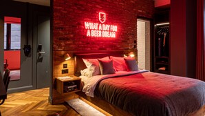 Habitació d'hotel moderna amb paret de maó, un cartell de neó que diu “What a day for a beer dream”, un llit de fusta, una tauleta de nit i una cadira vermella.