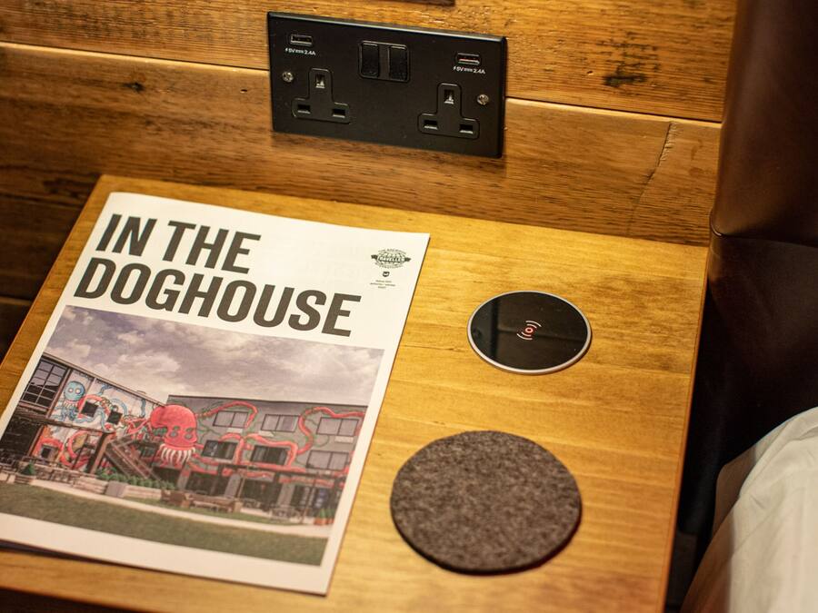 A wooden bedside table with a magazine titled "IN THE DOGHOUSE" and a circular object, possibly a speaker, on a hotel room.