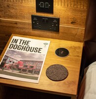Una mesita de noche de madera con una revista titulada “IN THE DOGHOUSE” y un objeto circular, posiblemente un altavoz, en una habitación de hotel.